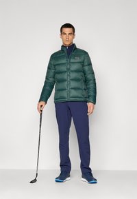 Green padded jacket with vertical quilted design, zip front, and branded patch. Worn with navy pants and dark shoes, holding a golf club.