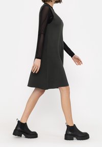 Robe sans manches vert foncé avec une coupe décontractée, superposée à un haut noir à manches longues en voile. Assortie à des bottes noires à talons épais.