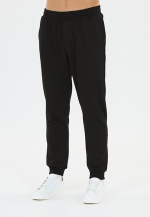 Mand iført sorte joggingbukser med elastisk talje og hvide sneakers, står på en hvid baggrund.