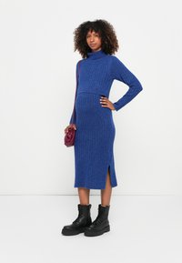 Blue ribbed knit dress with long sleeves and a high neckline, featuring side slits and paired with black lace-up boots and a purple bag.