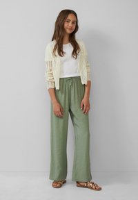 Cremefarbener Strickcardigan, weißes Hemd, hellgrüne weite Hose und braune Sandalen mit goldenen Akzenten, vor einem neutralen Hintergrund.
