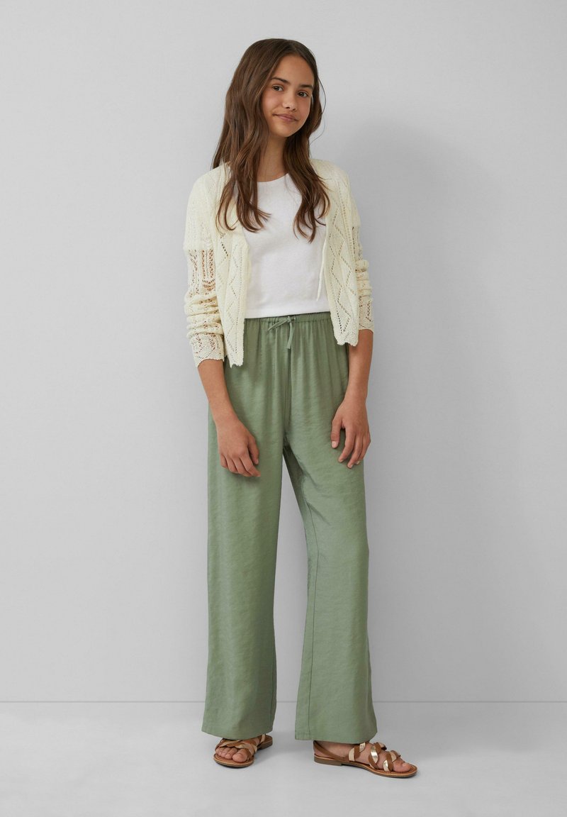 Cremefarbener Strickcardigan, weißes Hemd, hellgrüne weite Hose und braune Sandalen mit goldenen Akzenten, vor einem neutralen Hintergrund.