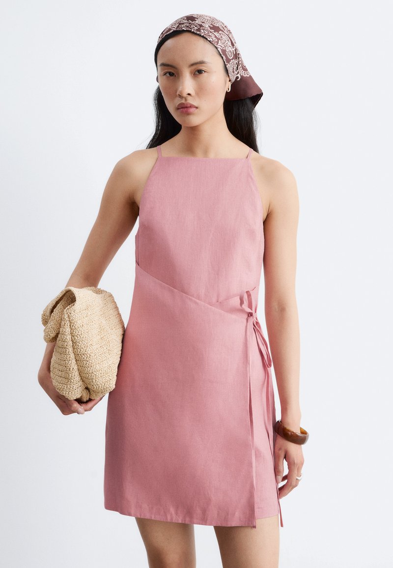 Femme portant une robe portefeuille rose sans manches, un foulard bordeaux à motifs, un bracelet en bois, tenant un sac tissé beige plié.
