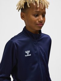 Jeune personne avec des dreadlocks blondes portant une veste zippée bleu marine avec le logo blanc hummel sur la poitrine, regardant légèrement vers la droite.