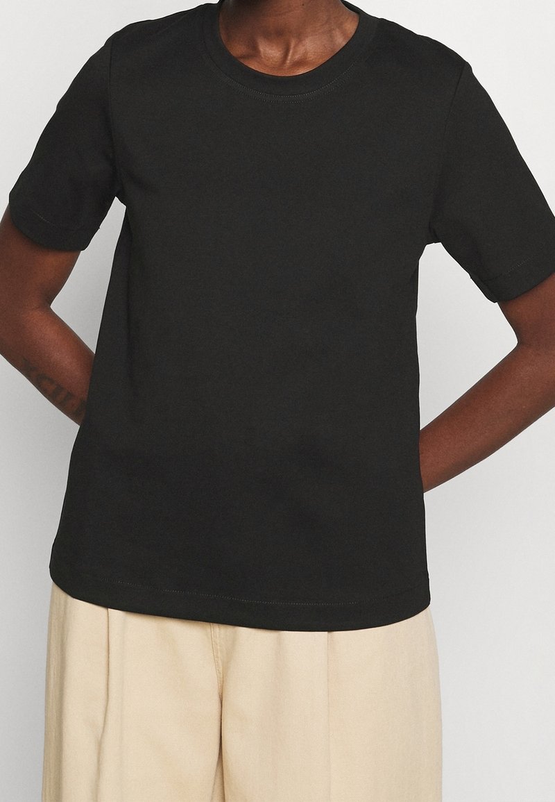 Personne portant un t-shirt noir uni à manches courtes et un pantalon beige ample, debout avec les mains dans le dos.