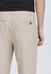 Beige kalhoty s texturovanou tkaninou, elastickým pasem a dvěma zadními kapsami, jedna má malou nášivku s logem. Košile je černá.