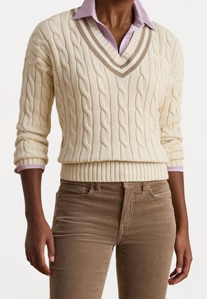 Personne portant un pull écru à torsades sur une chemise à col violet clair et un pantalon en velours côtelé marron, les mains détendues le long du corps.