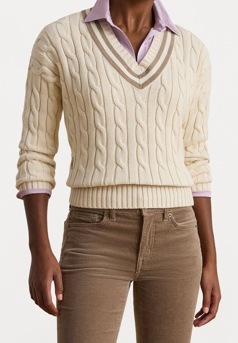 Personne portant un pull écru à torsades sur une chemise à col violet clair et un pantalon en velours côtelé marron, les mains détendues le long du corps.