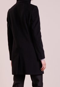 Sort uldfrakke med høj krave, lige silhuet og lange ærmer. Har en glat tekstur og slids bagpå for bevægelsesfrihed.