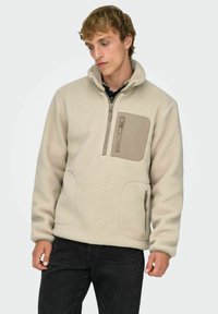 Kermansävyinen fleece-pulloveri, jossa on korkea kaulus, sivutaskut ja vetoketjulla suljettava rintatasku. Pinta on teksturoitu ja siinä on kontrastiväriset tan-värit.