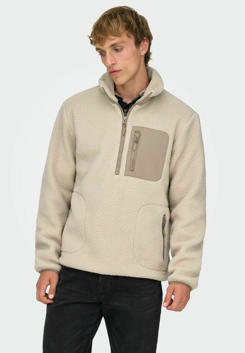 Kermansävyinen fleece-pulloveri, jossa on korkea kaulus, sivutaskut ja vetoketjulla suljettava rintatasku. Pinta on teksturoitu ja siinä on kontrastiväriset tan-värit.