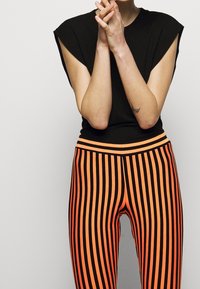 Svart topp med ärmar i form av kappa, tillsammans med orange och svarta randiga leggings. Outfiten har en figurnära design och ett brett elastiskt midjeband.