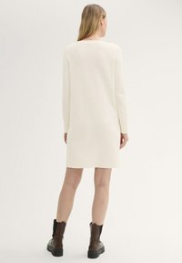 Weißes, gestricktes Sweaterkleid mit langen Ärmeln, geripptem Saum und geradem Schnitt, kombiniert mit braunen Knöchelstiefeln.