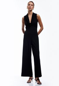 Schwarzer Jumpsuit mit einem Wasserfall-Ausschnitt, tailliertem Oberteil und weiten Beinen. Glatter Stoff mit drapiertem Design. Vertikale Linie betont die Silhouette.
