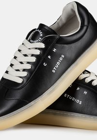 Baskets en cuir noir avec des lacets crème, ornées d'un texte imprimé en blanc "C P H STUDIOS." Semelle extérieure en caoutchouc semi-transparente avec un motif de traction.