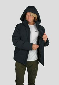 Homme portant une doudoune noire à capuche sur un t-shirt blanc et un pantalon cargo olive, fermeture éclair partiellement ouverte, ajustant la poche intérieure de la veste.