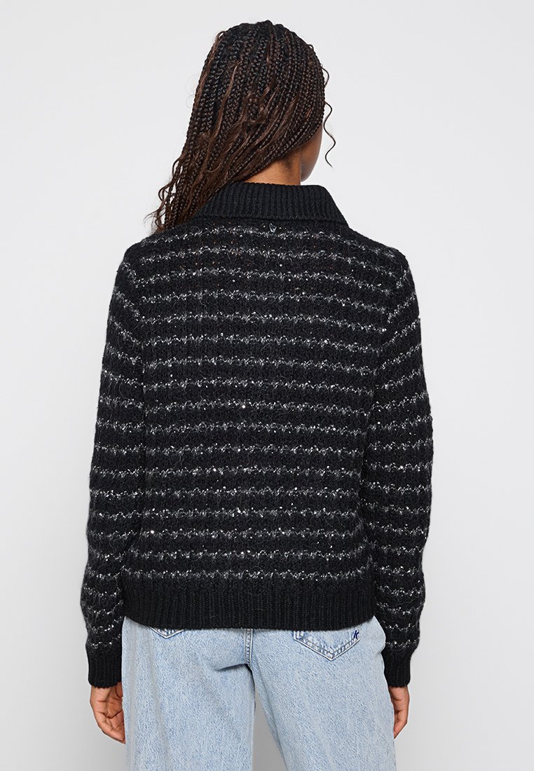 Vrouw met gevlochten haar, gekleed in een zwart en grijs gestreepte gebreide trui en lichte spijkerbroek, gezien van achteren tegen een effen achtergrond.