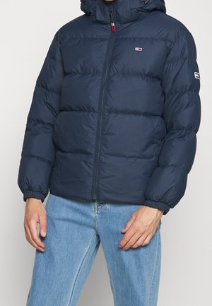 Homme portant une doudoune bleu marine avec capuche et un jean bleu clair, debout devant un fond uni clair.