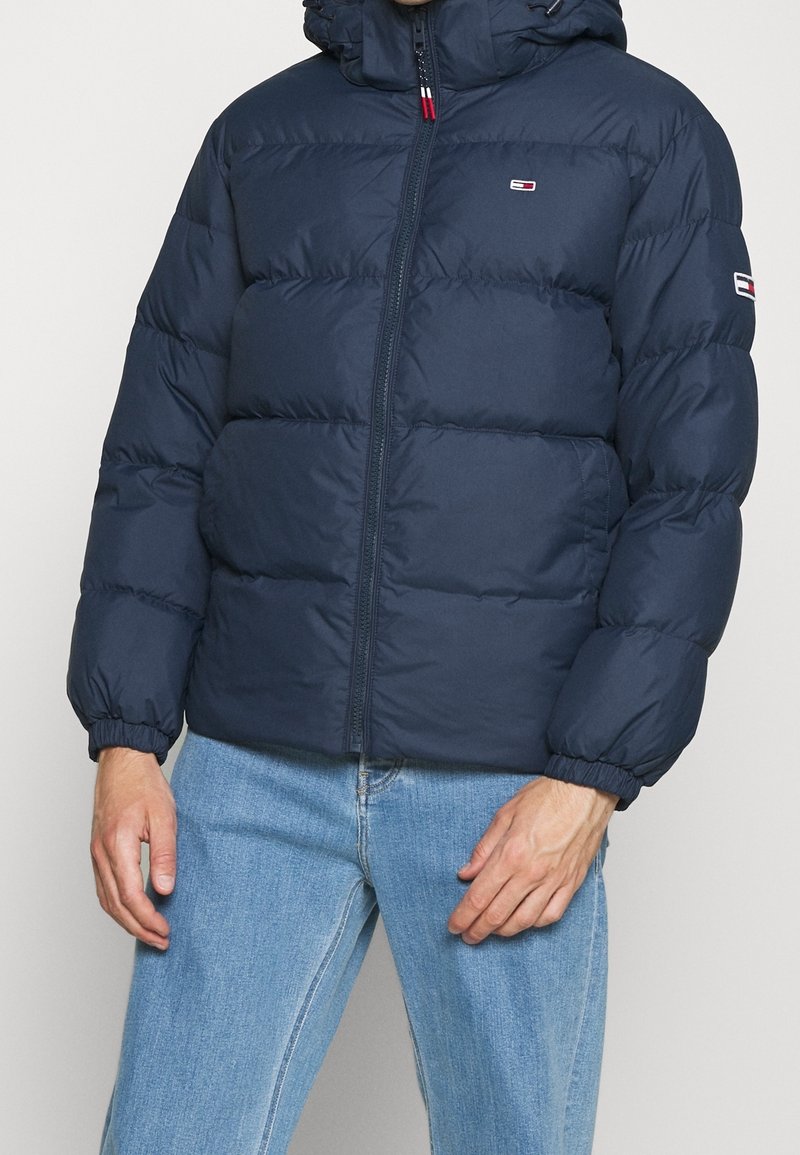 Homme portant une doudoune bleu marine avec capuche et un jean bleu clair, debout devant un fond uni clair.