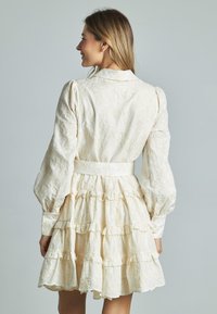 Helles cremefarbenes Kleid mit floralem Stickereien, langen Puffärmeln, tailliertem Schnitt und gestuften Rüschensaum. Knopfdetails an den Ärmelbündchen.