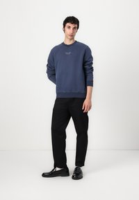 Felpa della marina, colletto a giro, polsini e orlo a costine, con dettagli testuali minimi. Abbinata a pantaloni neri e scarpe nere.