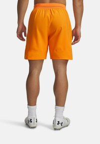 Asmuo stovintis su ryškiu oranžiniu sportiniu šortų, baltais Under Armour futbolo batais ir baltais trumpais kojinėmis, matomas iš nugaros.