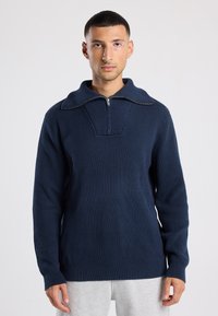 Man with short dark hair wearing a navy ribbed knit sweater with half-zip collar and light gray pants, standing against a plain background.