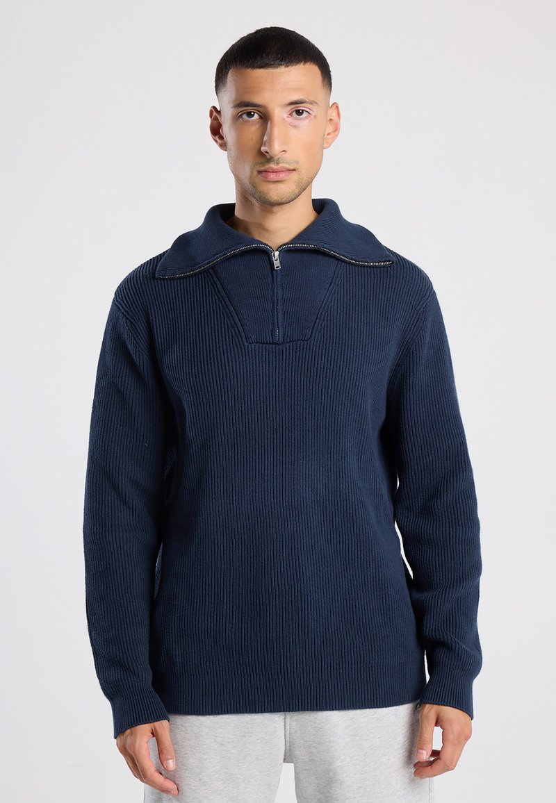 Man with short dark hair wearing a navy ribbed knit sweater with half-zip collar and light gray pants, standing against a plain background.