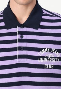 Stripet polo-skjorte i nyanser av lilla og marineblå, med krage, tre svarte knapper og brodert 'Tommy Jeans University Club'-logo.