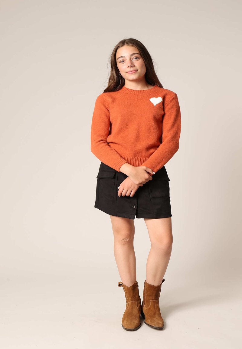 Un pull en tricot orange avec un détail en forme de cœur blanc, associé à une jupe noire. Des bottines en cuir marron complètent la tenue. Tissu texturé et design ajusté.