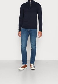 Granatowy sweter z zamkiem przy szyi i teksturowaną górną częścią, w połączeniu z niebieskimi, dopasowanymi jeansami i brązowo-białymi tenisówkami z płótna.