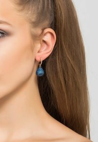 Pendientes de gota de cristal azul con forma de lágrima, acentuados por un aro en tono dorado. Los cristales muestran un detallado intrincado y brillan.