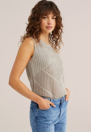 Vrouw met krullend bruin haar die een mouwloos lichtgrijs gebreid topje en blauwe spijkerbroek draagt, staand met handen in de zakken tegen een neutrale achtergrond.