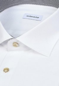 Gros plan sur le col d'une chemise blanche avec des boutons beiges et une étiquette de marque visible à l'intérieur portant la mention « seidensticker ».