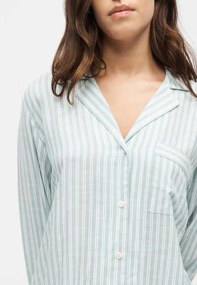 Femme portant une chemise à rayures verticales bleu clair et blanche avec des boutons et une poche, montrée du cou jusqu'au milieu du torse sur un fond uni.