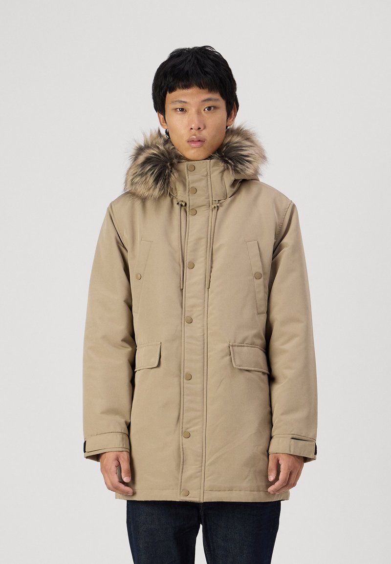 Parka di colore beige con cappuccio foderato in pelliccia, caratterizzata da bottoni a pressione e due tasche frontali. Realizzata in un tessuto liscio e resistente.
