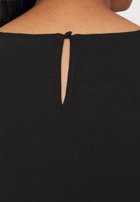 Svart tygblus med rund halsringning och en liten nyckelhålssmyckning bak som hålls ihop med en svart knapp. Mjuk textur och figurnära design.