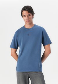 Camiseta de algodón azul con escote redondo y mangas cortas. Presenta un logotipo sutil en rojo oscuro en el frontal. Corte relajado.