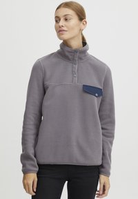 Pullover in pile grigio con colletto alto, dotato di taschino blu sul petto, patta con tre bottoni e vestibilità comoda. Texture morbida in tutta la superficie.