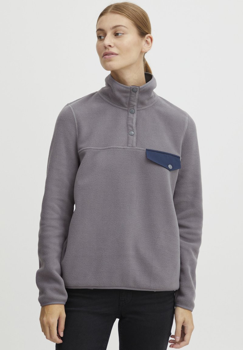 Pullover in pile grigio con colletto alto, dotato di taschino blu sul petto, patta con tre bottoni e vestibilità comoda. Texture morbida in tutta la superficie.