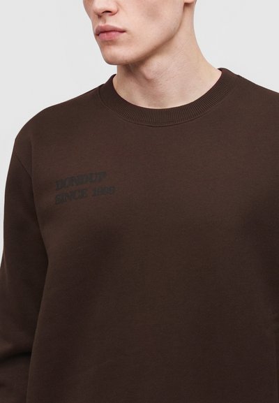 Jeune homme portant un sweat à col rond marron foncé avec un texte noir pâle indiquant « "DIALOGUE SINCE 1980" » sur le côté gauche de la poitrine.
