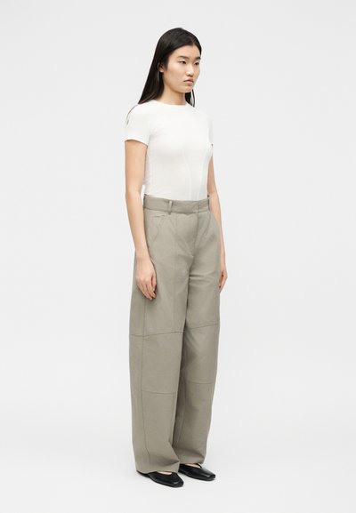 Jeune femme debout portant un haut blanc cintré à manches courtes, un pantalon beige à jambes larges, et des ballerines pointues noires devant un fond clair uni.