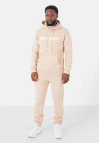 Ensemble de sweat à capuche rose clair et de joggeurs en tissu doux, avec une poche kangourou et un logo imprimé blanc. Porté avec des baskets blanches.