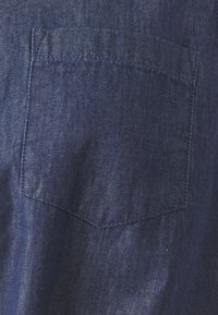 Il tessuto di jeans presenta una piccola tasca a punta. Il materiale è di colore blu scuro con una sottile texture, e le cuciture sono visibili lungo i bordi.