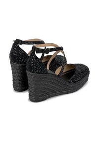 Sandali con tacco a cuneo neri con suola intrecciata, texture in suede, adornati con borchie nere, presentano cinturini incrociati e una cinghia alla caviglia regolabile.