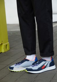 Scarpe sportive con pannelli multicolori in tonalità di blu, grigio e arancione, dotate di superfici texture e una suola a zigzag. Pantaloni neri e calzini bianchi.