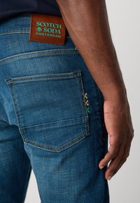Los vaqueros de mezclilla en un tono azul medio presentan una etiqueta de cuero con "Scotch & Soda Amsterdam". Destacan costuras de colores en el bolsillo.