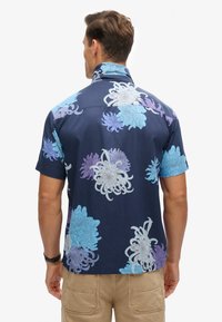 Camisa de manga corta en azul marino oscuro con grandes patrones florales en azul claro, lavanda y blanco. Tejido suave, cuello estándar, corte relajado.