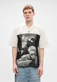 Πουκάμισο με κουμπιά μπροστά που έχει λευκό πάνω μέρος και μεγάλο μονόχρωμο γραφικό σχέδιο με τρεις χαρακτήρες στο κάτω μισό; άνετη εφαρμογή και κοντά μανίκια.