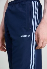 Tmavě modré sportovní kalhoty z měkké látky s světle modrými pruhy po stranách a vyšívaným bílým logem Adidas na levém stehně.